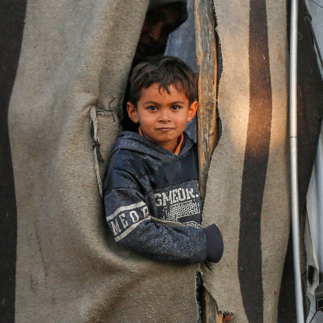 A young Palestine boy peeks through a torn blanket used as a door in a refugee camp shelter. His quiet expression reflects the resilience of children living in displacement and hardship. The image powerfully symbolizes the daily struggles faced by Palestine refugee families.