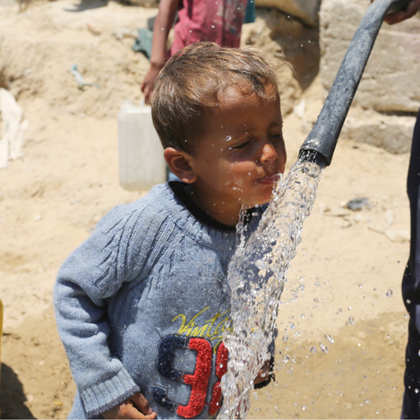 A young Palestine Refugee boy drinks water directly from a hose in a refugee camp, highlighting the urgent need for clean and accessible water in Gaza. The image captures a moment of relief amid ongoing hardship and water scarcity faced by displaced communities.