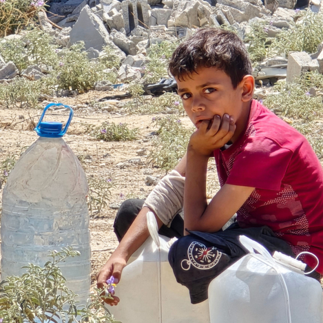 A young boy in Gaza sits beside water containers amid rubble, highlighting the urgent humanitarian crisis and the struggle of Palestine Refugees for access to clean water and basic needs.