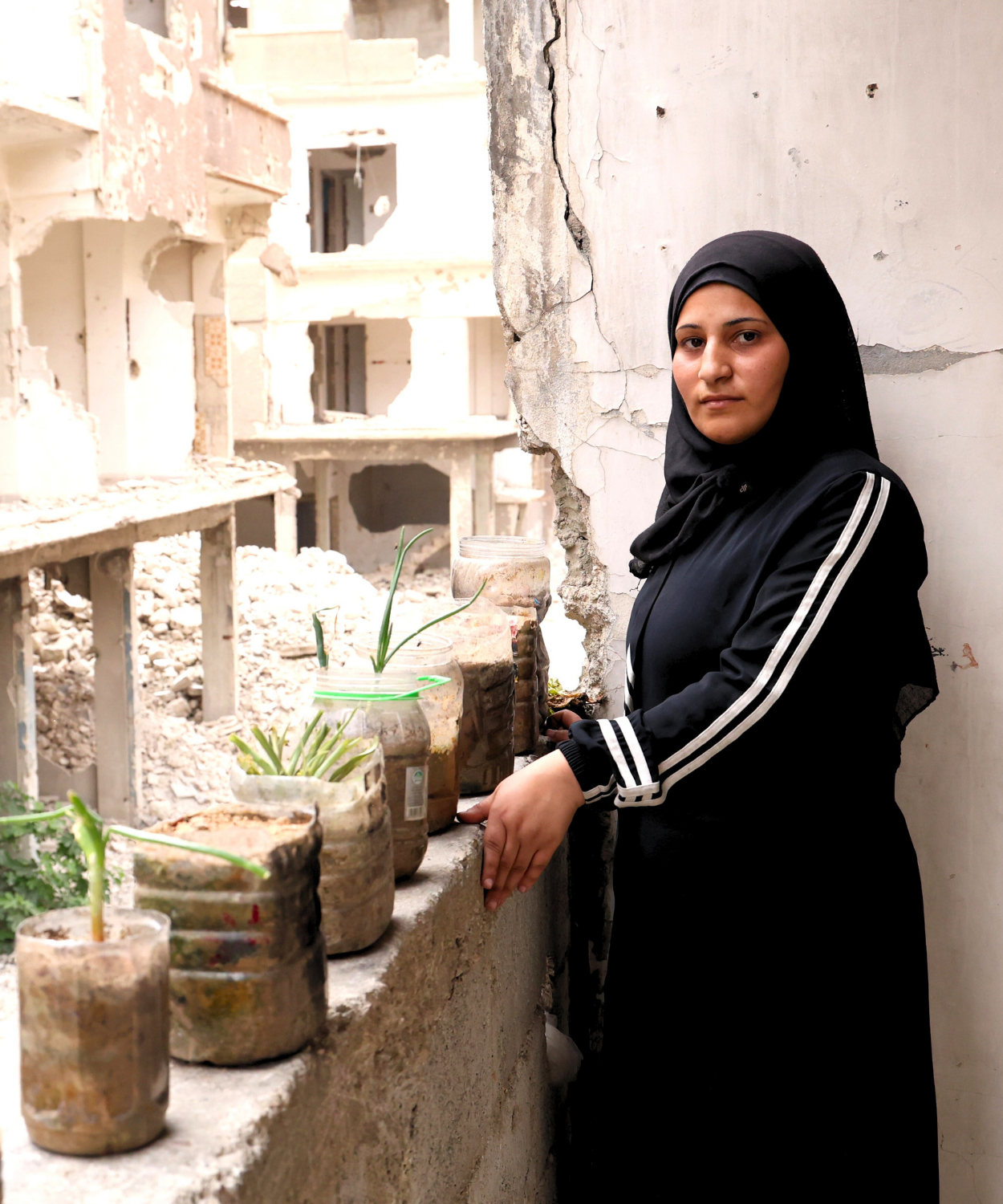A Palestine Refugee woman in Syria stands on the balcony of her damaged home with freshly planted greenery, symbolizing hope and rebuilding amidst destruction.