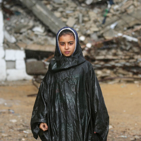 A young Palestinian girl shelters herself from the rain using a plastic bag, highlighting the urgent humanitarian needs faced by refugee families supported through UNRWA Zakat.