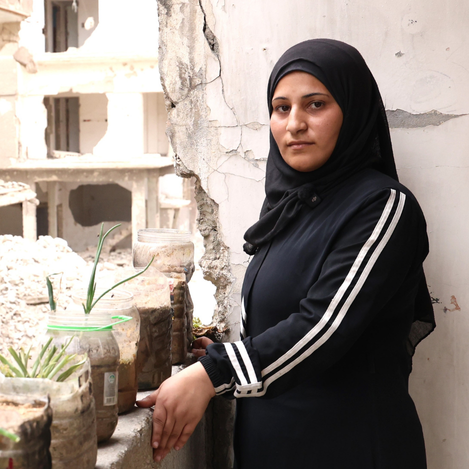 A Palestine Refugee woman in Syria stands beside her damaged home, highlighting resilience, displacement, and the urgent need for humanitarian support.
