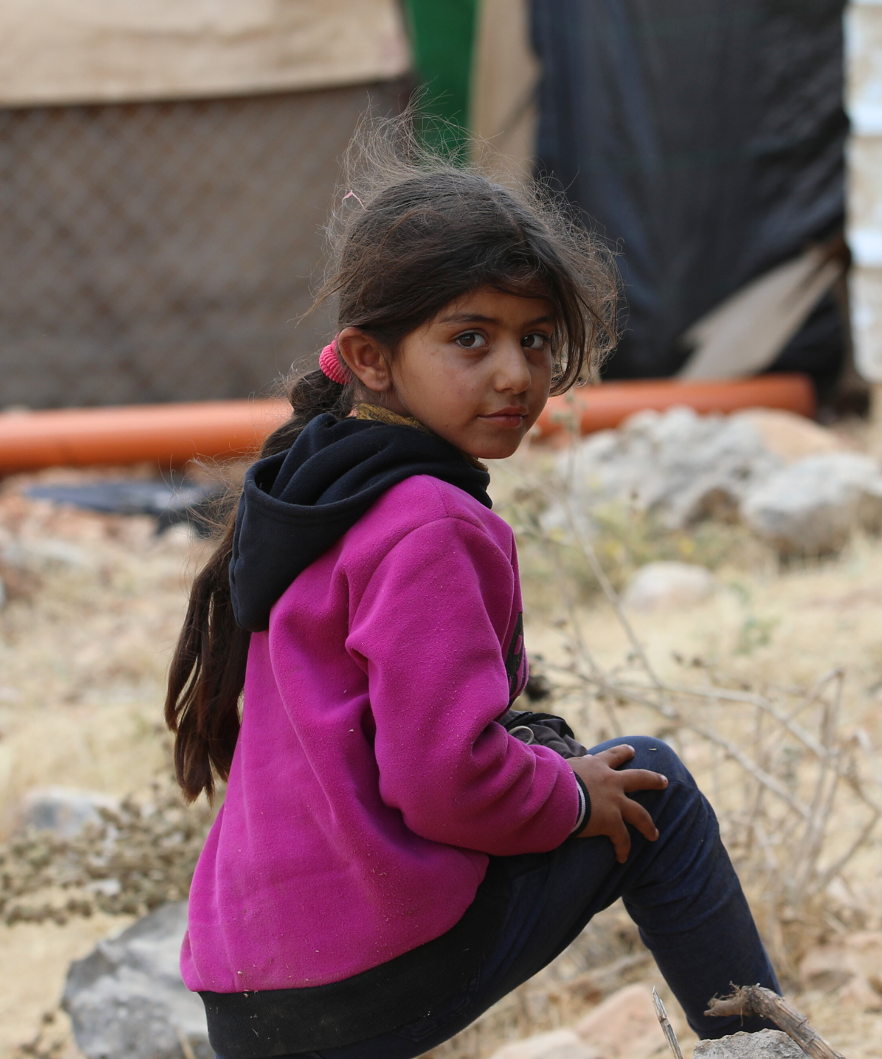 Portrait of a young Palestinian refugee girl in the West Bank, East Jerusalem area, sitting among rubble and debris. The child wears a bright pink sweatshirt and black pants, looking directly at the camera with a poignant expression. Documentary photography capturing the reality of Palestinian refugee children living in difficult conditions in the occupied West Bank and East Jerusalem, highlighting the impact of displacement on young lives and the resilience of Palestinian youth.