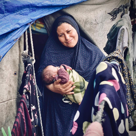 UNRWA Palestine Refugee family: adult holding an infant inside a tent shelter in a displacement camp.