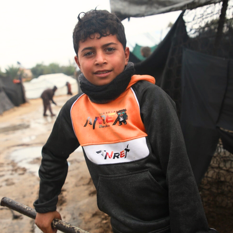 A child standing in rain near rubble in Gaza, highlighting humanitarian crisis