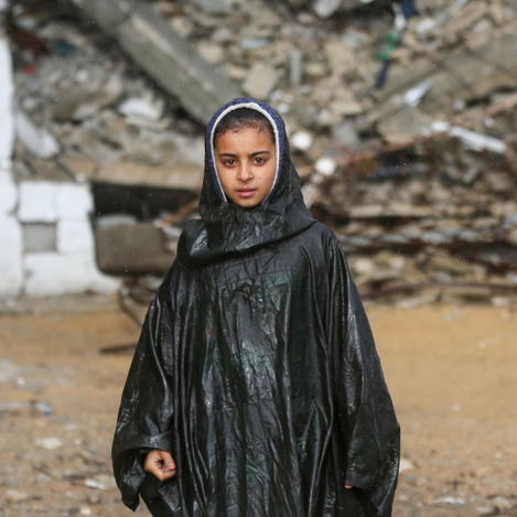 A young Palestinian girl shelters herself from the rain using a plastic bag, highlighting the urgent humanitarian needs faced by refugee families supported through UNRWA Zakat.